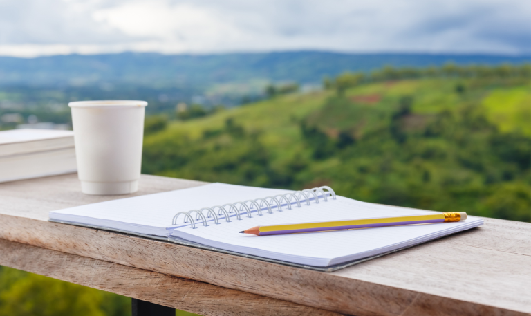 Cup, notebook and pencil on a wooden shelf with countryside behind them