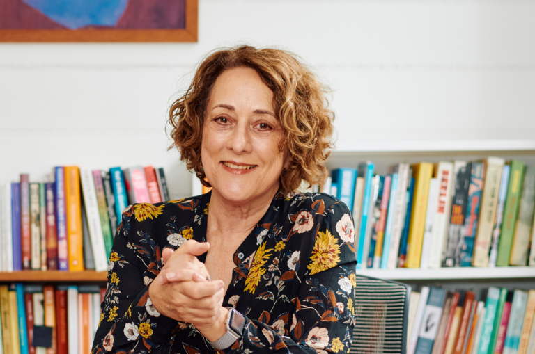 A woman sits behind a desk with her hands clasped in front of her. There are bookshelves filled with books behind her.