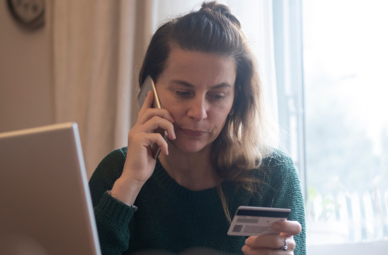 Woman sitting in front of her computer, on her mobile reading out the details of her credit card.